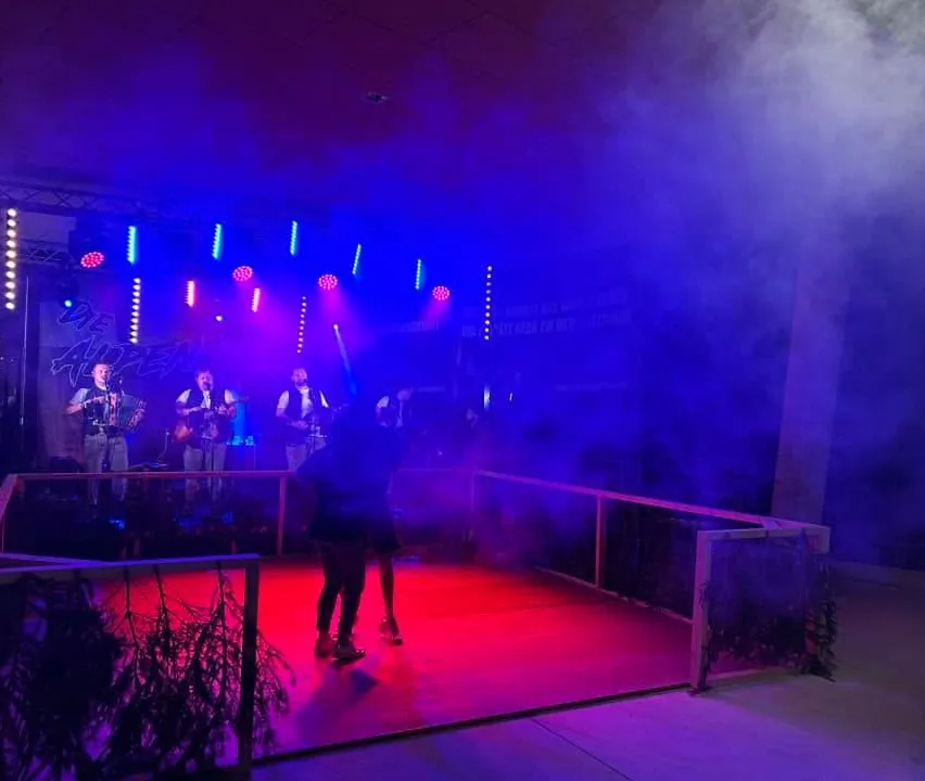 Eine Band spielt auf einer Bühne mit roter und blauer Beleuchtung. Die Bühne ist von einem Geländer umgeben, und ein Paar tanzt im Vordergrund.