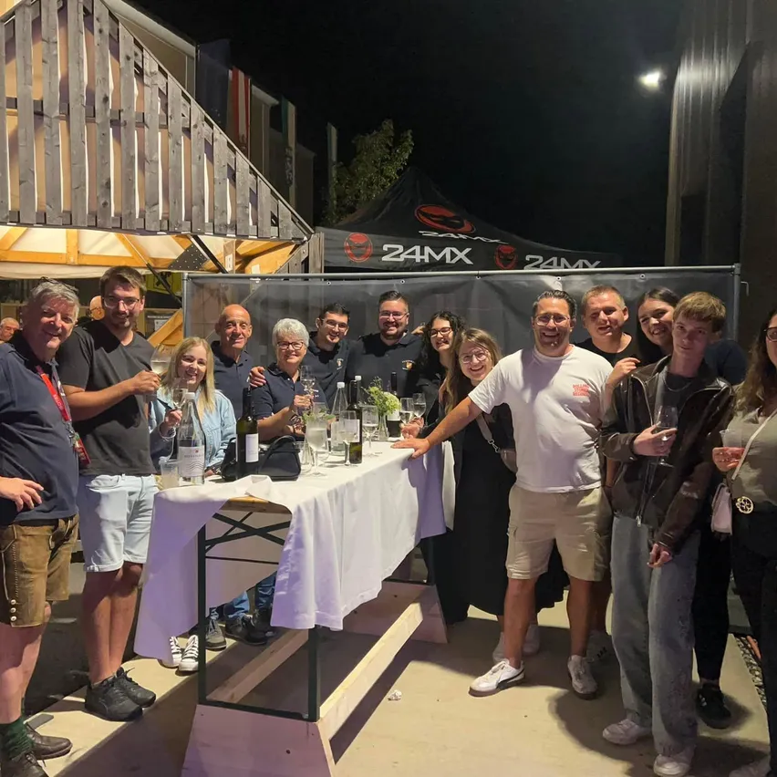 Eine Gruppe von Menschen steht um einen Tisch mit Getränken und Gläsern herum, lächelt und posiert für ein Foto bei Nacht.