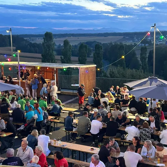 Eine große Gruppe von Menschen versammelt sich in einem Outdoor-Essbereich mit langen Holztischen und Bänken unter bunten Regenschirmen. Die Umgebung ist von Bäumen und einer malerischen Aussicht auf die Landschaft umgeben.
