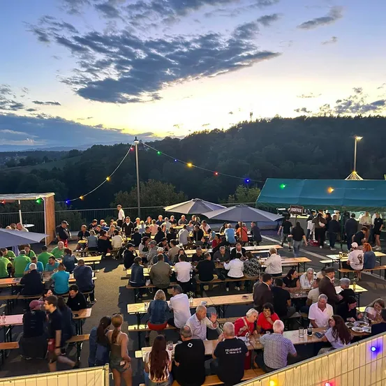 Eine lebhafte Outdoor-Essensbereich mit vielen Menschen an Tischen, bunten Lichtern und einer malerischen Kulisse von Hügeln und Sonnenuntergang.