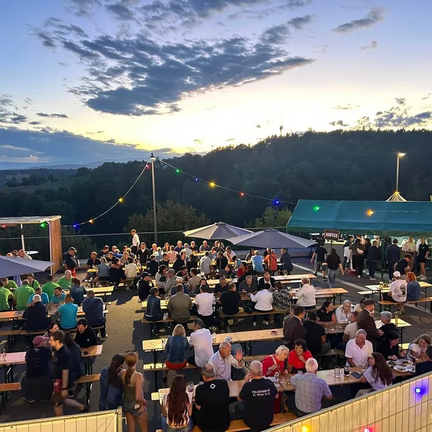 Eine lebhafte Outdoor-Essensbereich mit vielen Menschen an Tischen, bunten Lichtern und einer malerischen Kulisse von Hügeln und Sonnenuntergang.