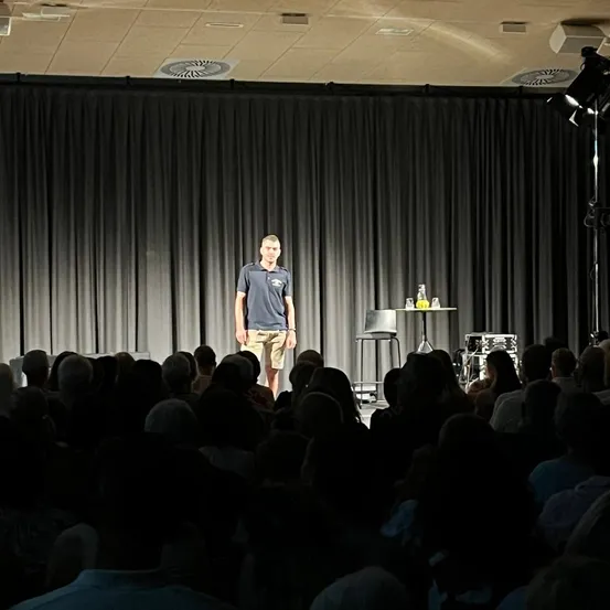 Ein Mann steht auf der Bühne vor einem sitzenden Publikum, mit einem Tisch, auf dem Flaschen stehen.