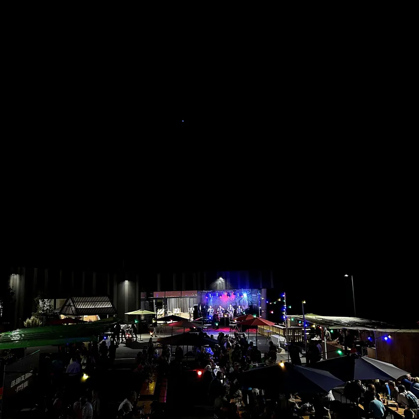 Ein lebhaftes Nachtmarkt mit einer Bühnenshow unter einem dunklen Himmel, beleuchtet von bunten Lichtern und besucht von einer Menge.