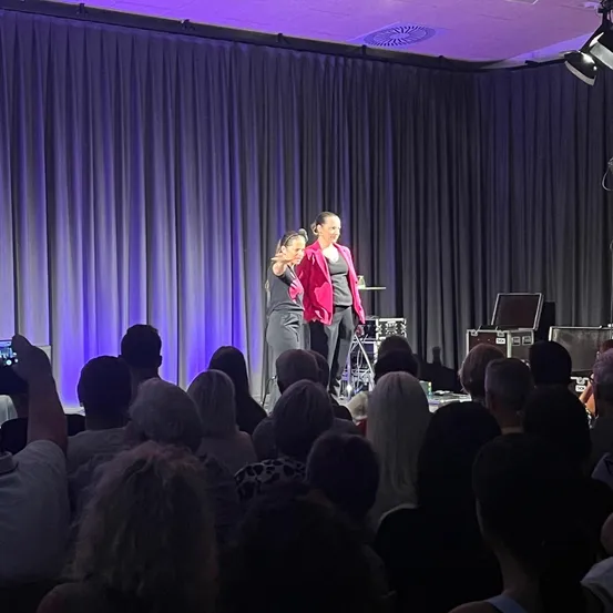 Zwei Frauen stehen auf der Bühne, eine in einem rosa Blazer, und sprechen vor einem Publikum in einem schwach beleuchteten Raum.
