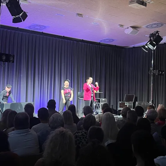 Drei Darsteller auf der Bühne mit lila Beleuchtung, eine Frau in Pink spricht, während die anderen zusehen. Das Publikum sitzt davor.