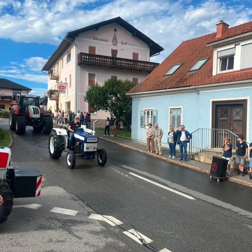 Ein Traktor-Parade auf einer Straße mit Zuschauern, ein Traktor führt mit einem Fahrer, und Gebäude auf beiden Seiten.