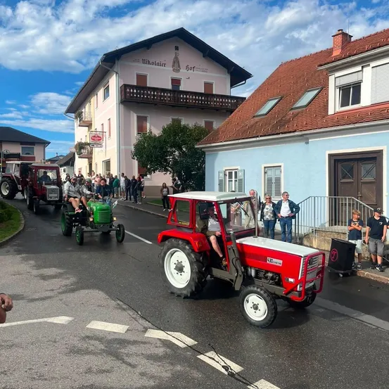 Ein roter Traktor führt eine Parade von Oldtimer-Traktoren die Straße hinunter, mit Zuschauern und Gebäuden im Hintergrund.