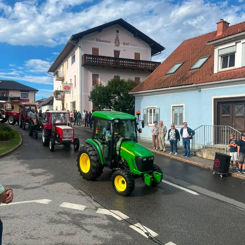 Ein Umzug von Oldtimer-Traktoren auf einer Straße mit Zuschauern und Gebäuden im Hintergrund.