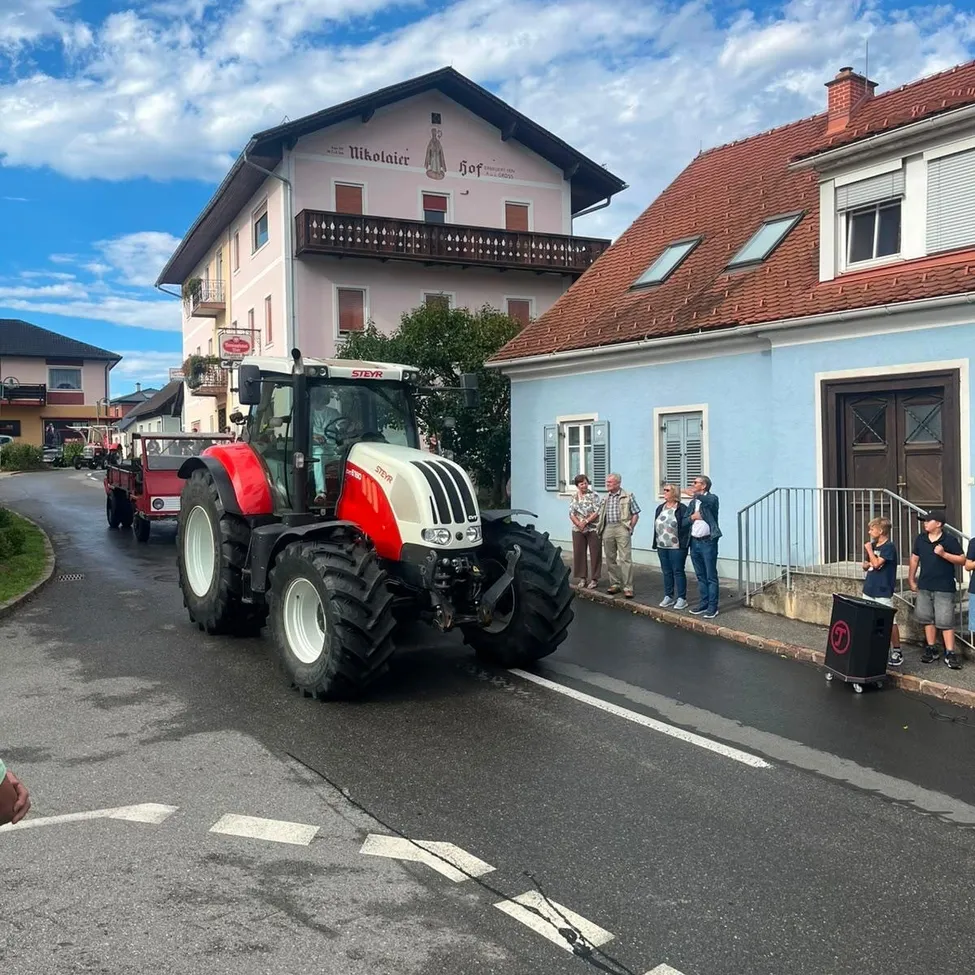 Ein Traktor fährt die Straße entlang, während Zuschauer auf dem Bürgersteig stehen. Gebäude säumen die Straße, eines mit Balkon.