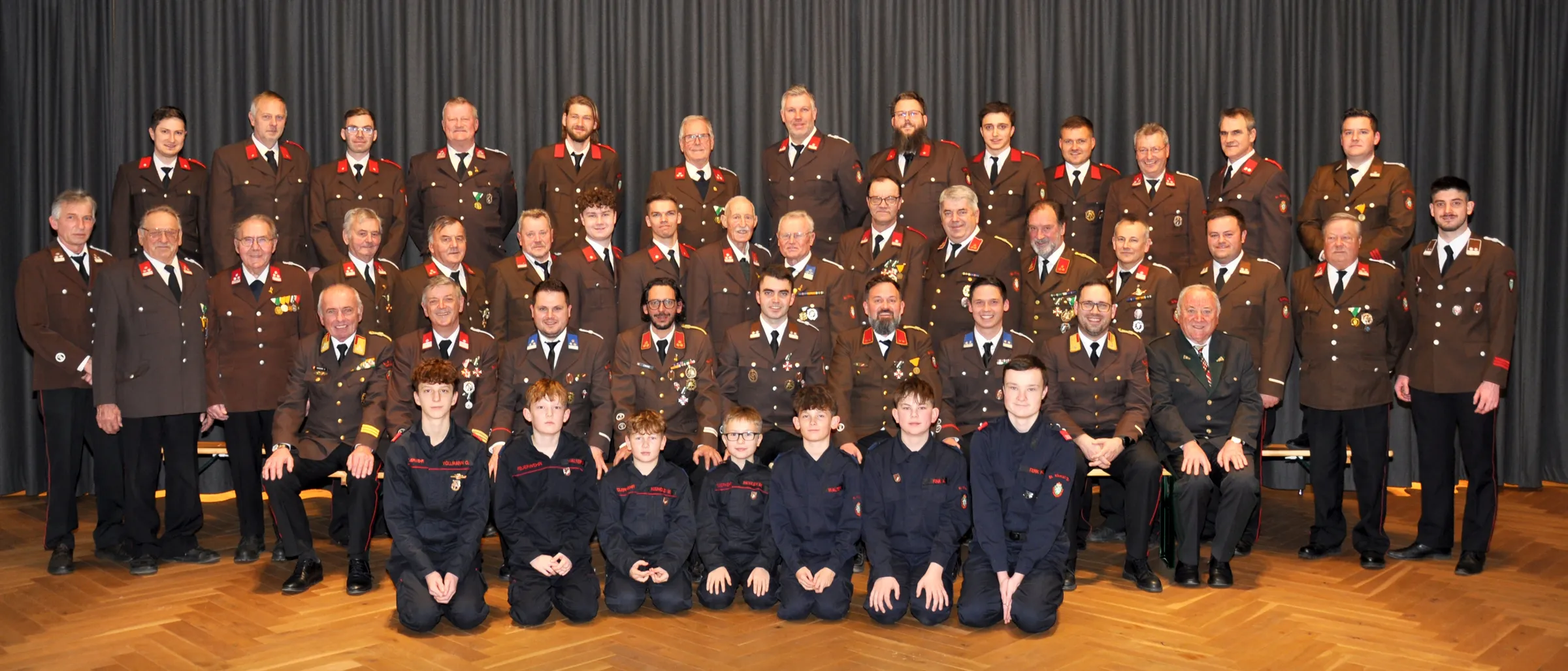 Eine Gruppe von Militäroffizieren und Kadetten posiert für ein Foto. Die Offiziere tragen Uniformen mit Medaillen, während die Kadetten schwarze Uniformen tragen. Sie alle lächeln und schauen in die Kamera.