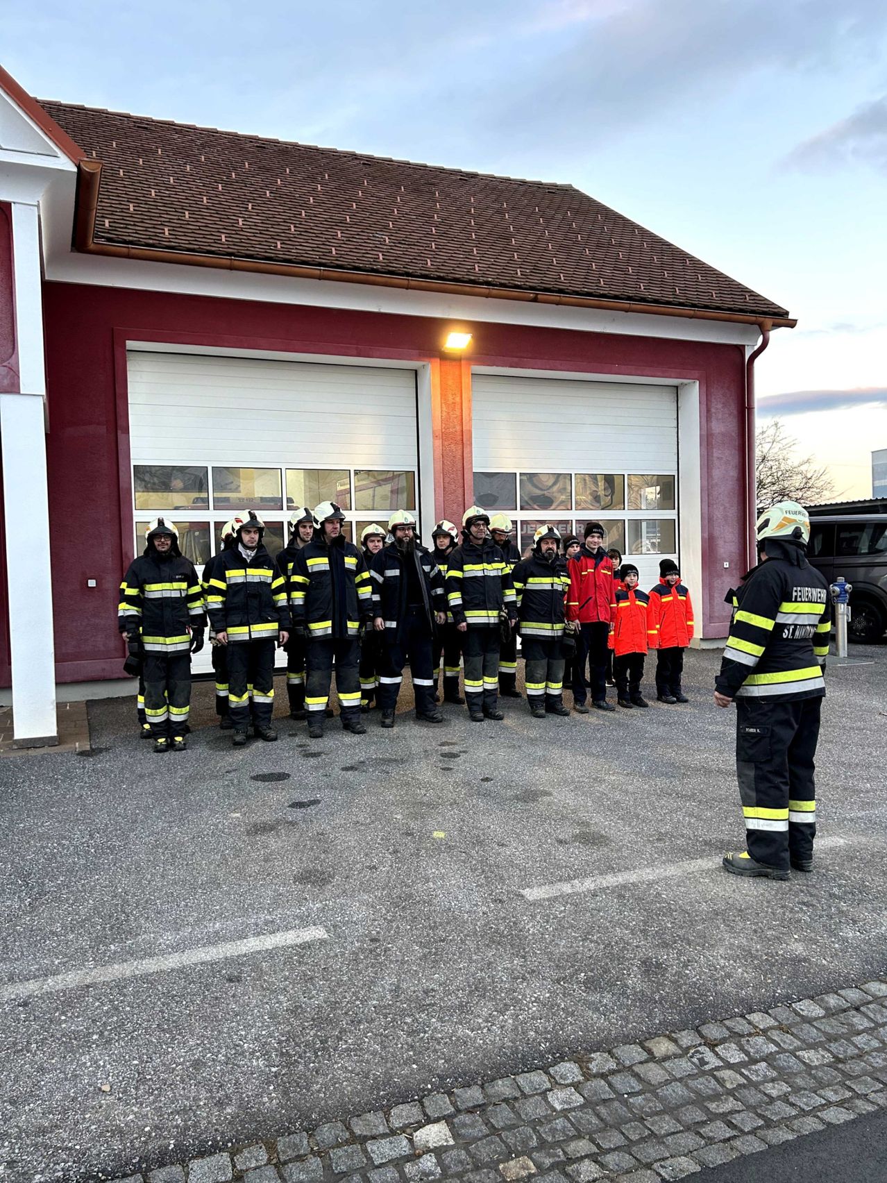 Eine Gruppe Feuerwehrleute steht in Uniform vor einer Feuerwache. Einige Kinder stehen in der Nähe, und ein Feuerwehrmann spricht sie an.