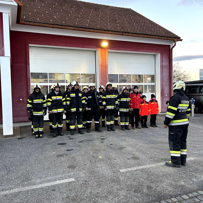 Eine Gruppe Feuerwehrleute steht in Uniform vor einer Feuerwache. Einige Kinder stehen in der Nähe, und ein Feuerwehrmann spricht sie an.