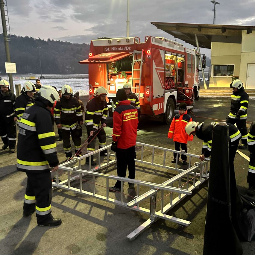 Eine Gruppe von Feuerwehrleuten ist draußen und bereitet ein Metallgerüst vor, während ein Kind zusieht. Ein roter Feuerwehrwagen ist in der Nähe geparkt.