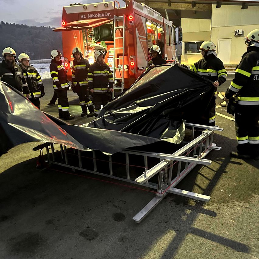 Eine Gruppe Feuerwehrleute in Uniform steht neben einem Feuerwehrwagen mit einem abgedeckten Krankentragen auf einem Metallrahmen.
