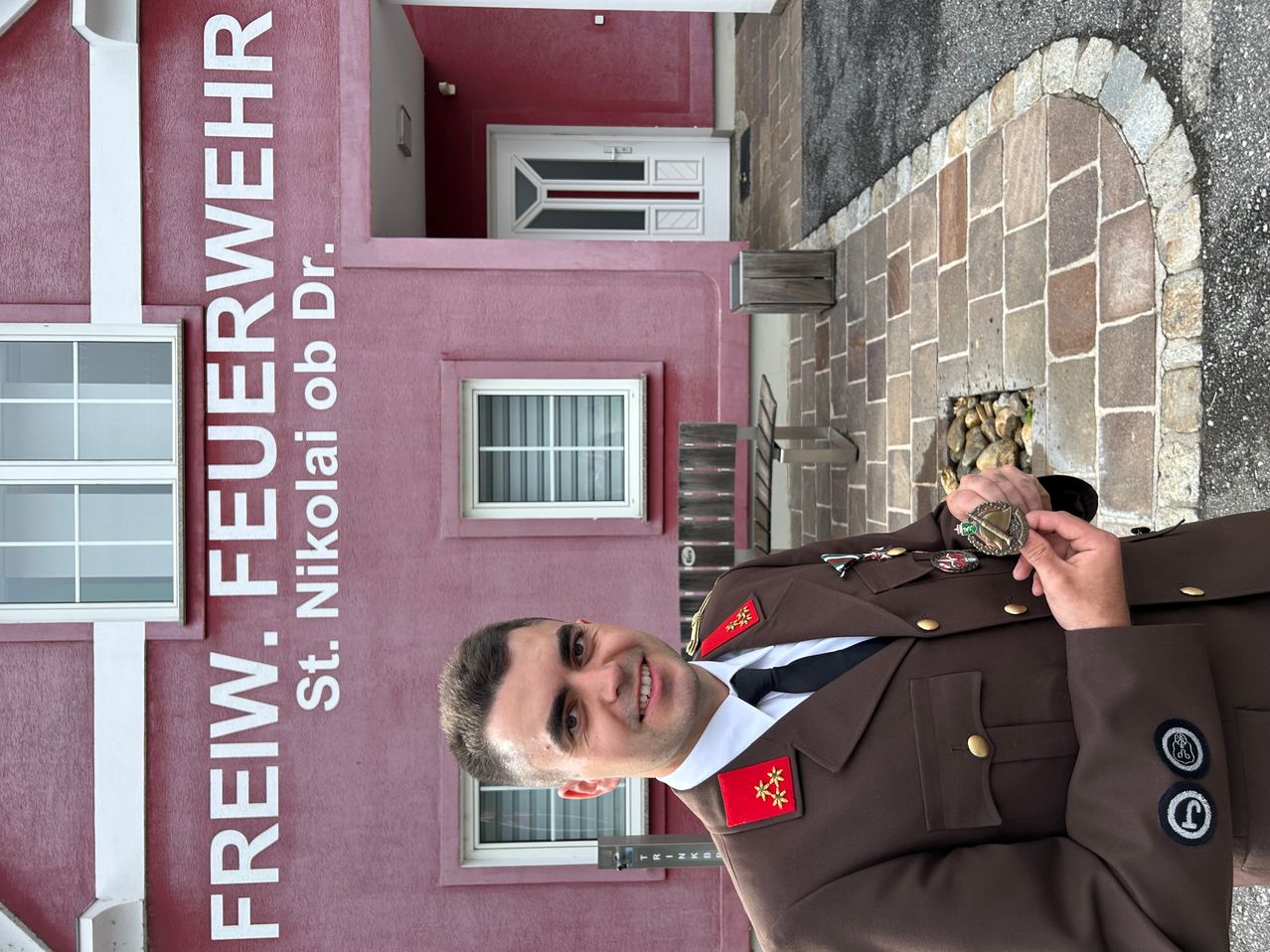 Ein lächelnder Mann in Uniform steht vor einem roten Gebäude mit der Aufschrift 'FREIW. FEURWEHR St. Nikolai ob Dr.'.