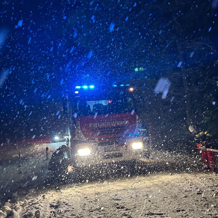 Ein Feuerwehrmann sägt mit einer Kettensäge durch den Schnee auf einer Straße, während ein Feuerwehrwagen vorbeifährt.