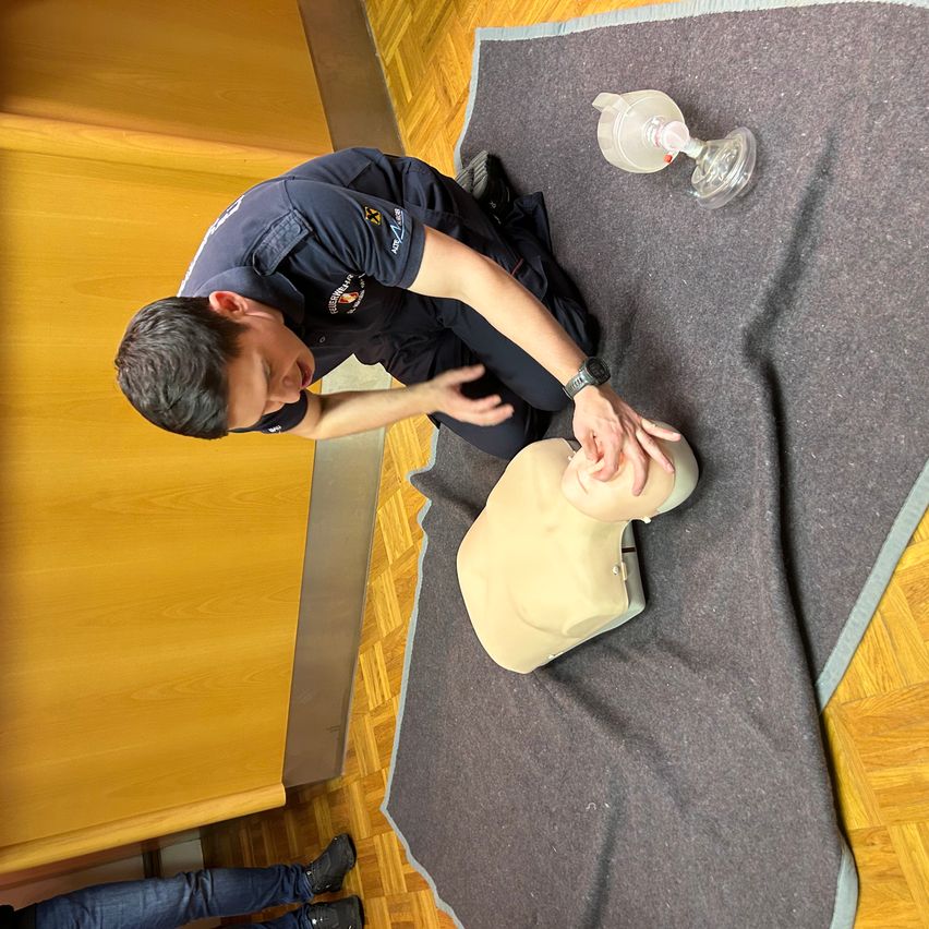 Ein Mann in einer dunkelblauen Uniform führt eine Herzdruckmassage an einer Puppe auf einem grauen Teppich durch, mit einem Wiederbelebungsgerät in der Nähe. Der Ort scheint ein Innenbereich mit Holzboden zu sein.