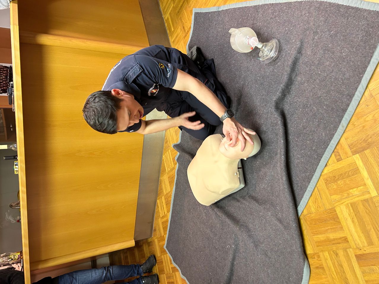 Ein Mann in einer dunkelblauen Uniform führt eine Herzdruckmassage an einer Puppe auf einem grauen Teppich durch, mit einem Wiederbelebungsgerät in der Nähe. Der Ort scheint ein Innenbereich mit Holzboden zu sein.