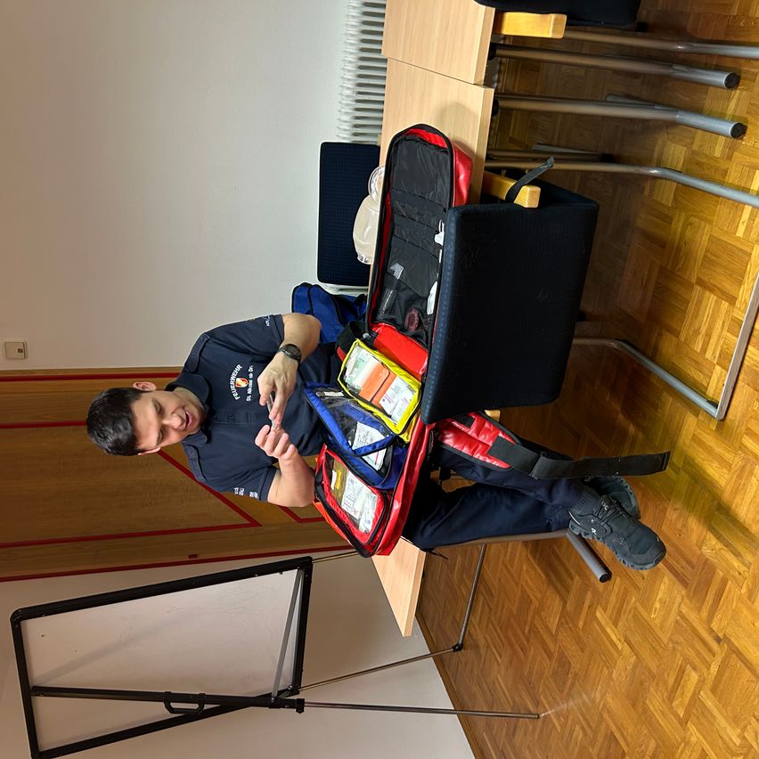 Ein Mann sitzt mit verschränkten Beinen auf einem Stuhl, hält eine Erste-Hilfe-Ausrüstung und lächelt. Er befindet sich wahrscheinlich in einem Klassenzimmer.