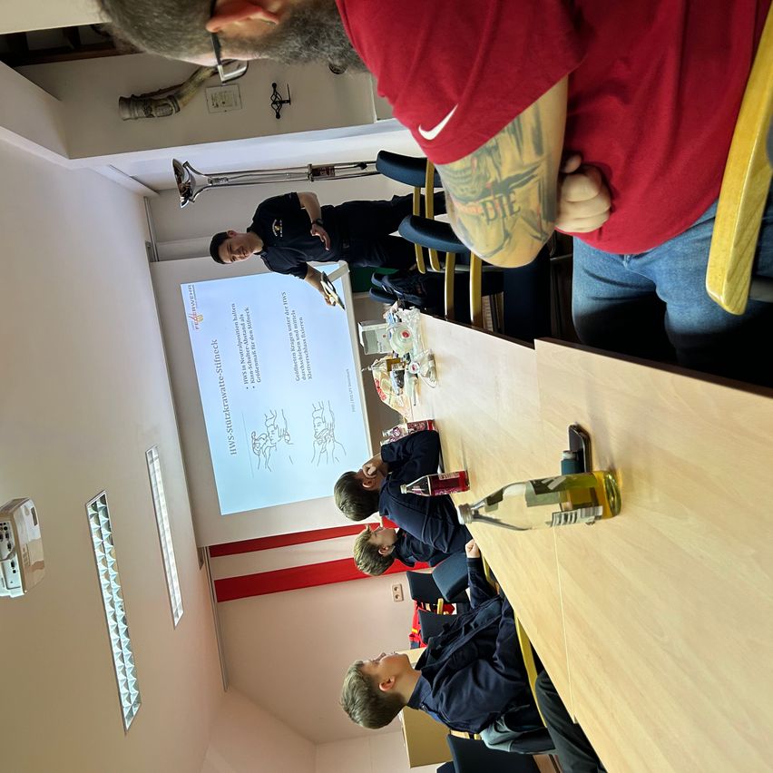 Eine Gruppe von Menschen sitzt in einem Klassenzimmer um einen Tisch. Ein Mann steht vor einer Projektionsleinwand.