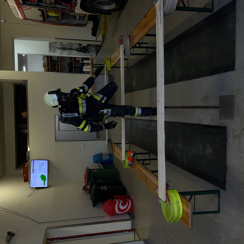 Ein Feuerwehrmann schwebt in der Luft und trägt eine vollständige Schutzausrüstung. Der Raum ist mit Trainingsgeräten, einem Monitor und einem im Hintergrund sichtbaren Feuerwehrwagen ausgestattet.