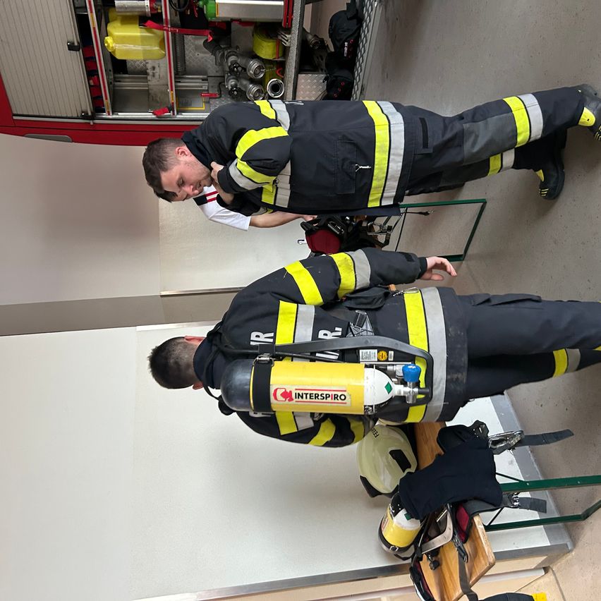 Zwei Feuerwehrleute in Schutzanzügen, einer kniend, inspizieren Ausrüstung, der andere beugt sich vor. Die Szene beinhaltet einen Feuerlöscher und einen Helm auf einem Holztisch.