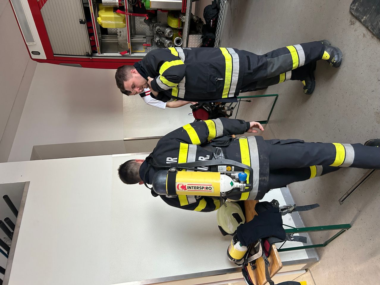 Zwei Feuerwehrleute in Schutzanzügen, einer kniend, inspizieren Ausrüstung, der andere beugt sich vor. Die Szene beinhaltet einen Feuerlöscher und einen Helm auf einem Holztisch.
