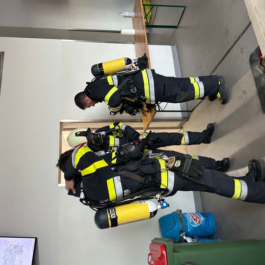 Drei Feuerwehrleute in Schutzkleidung sind in einem Raum. Einer sitzt, ein anderer kniet und der dritte steht. Ein Feuerlöscher und ein Monitor sind in der Nähe.