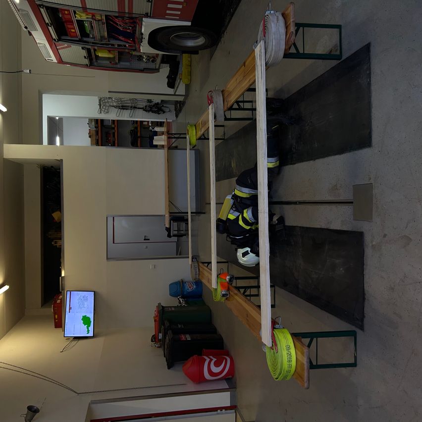 Eine Draufsicht eines Raumes mit einem Feuerwehrmann in voller Ausrüstung. Ein Monitor und Feuerlöscher sind sichtbar. Ein Feuerwehrauto ist geparkt, und der Raum ist mit verschiedenen Werkzeugen ausgestattet.