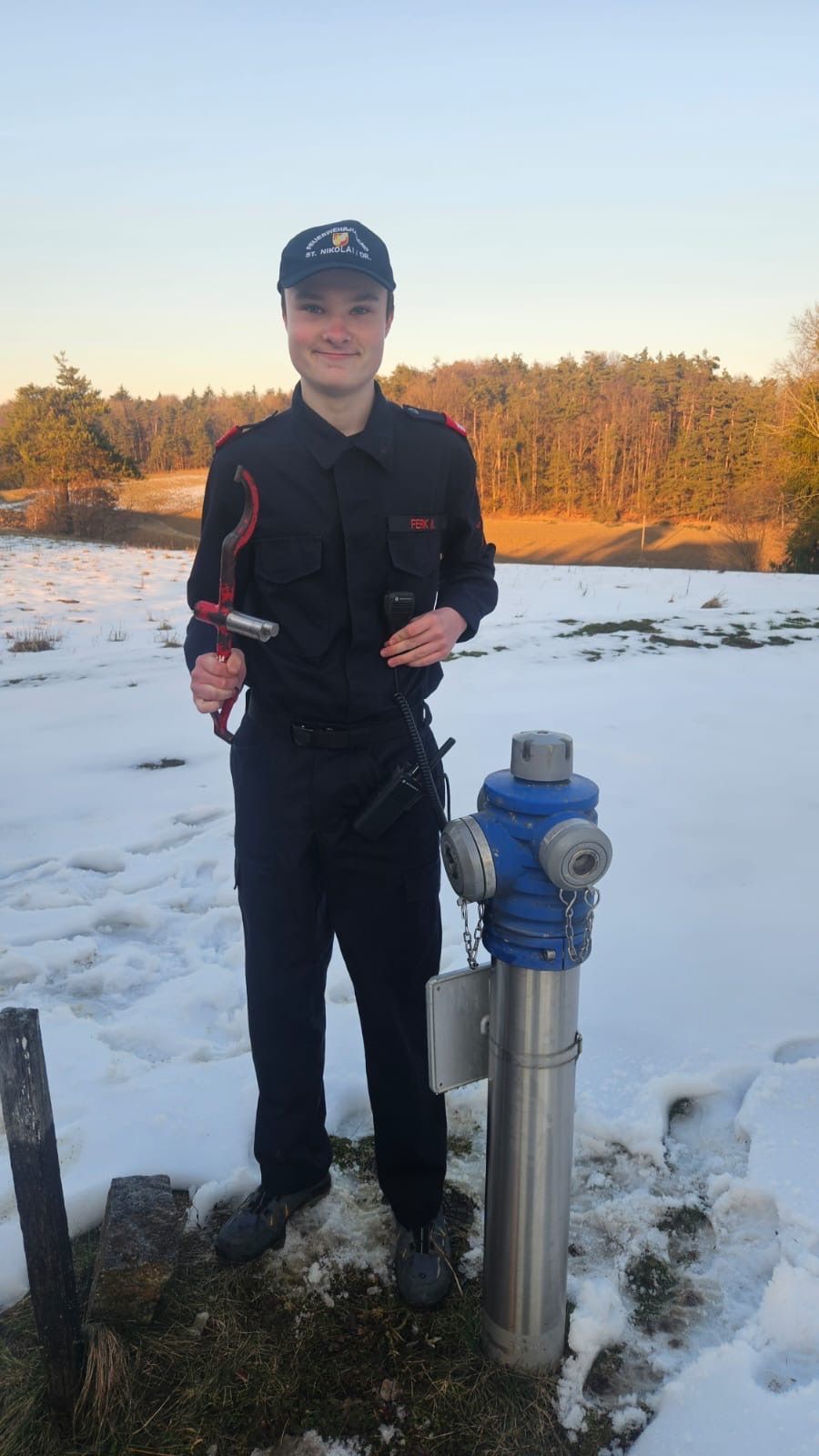 Eine Person in Uniform steht im Schnee neben einem Feuerhydranten, hält ein Werkzeug und ein Walkie-Talkie.