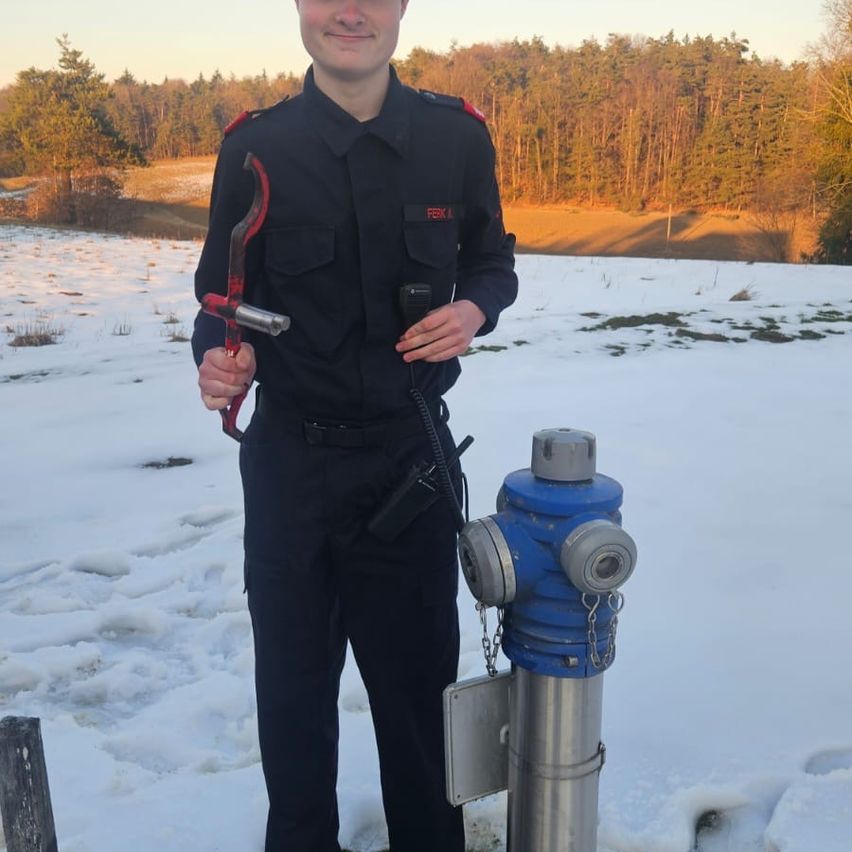 Eine Person in Uniform steht im Schnee neben einem Feuerhydranten, hält ein Werkzeug und ein Walkie-Talkie.