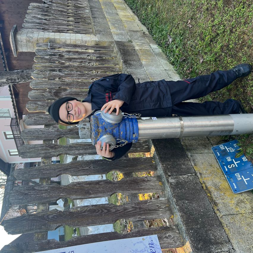 Ein junger Junge in einer schwarzen Jacke und einer Brille liegt gegen einen Holzzaun und hält eine rostige blaue und silberne Rohr. Hinter ihm befindet sich ein Gebäude mit Fenstern und einem Schild mit Zahlen.