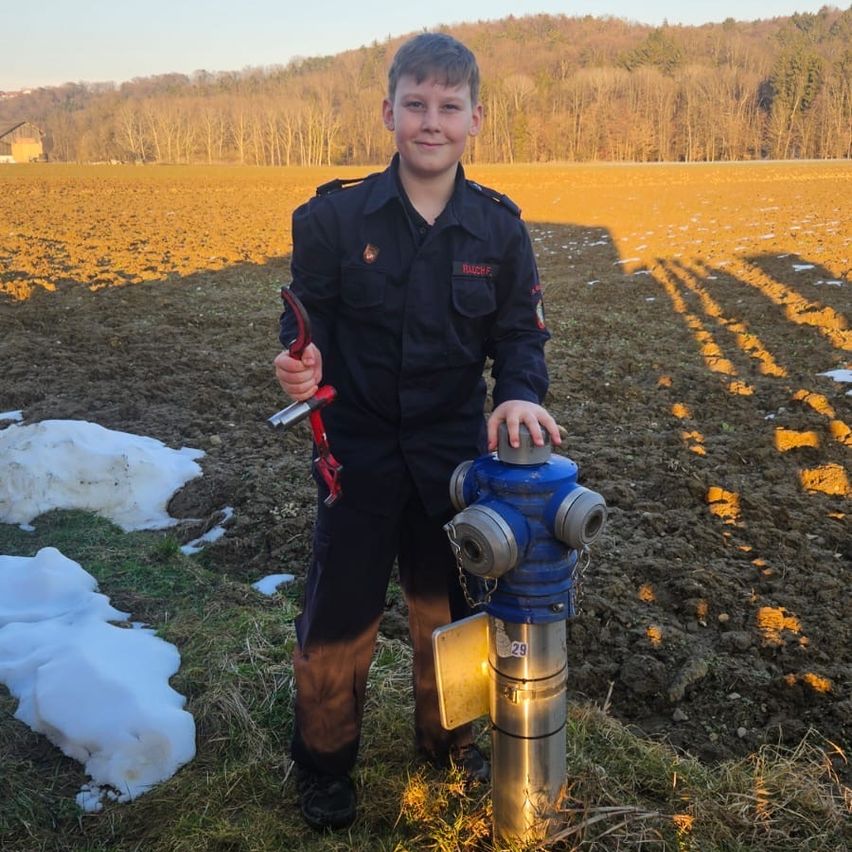Ein Junge in Uniform posiert mit einem Feuerhydranten auf einem schneebedeckten Feld, Schatten auf dem Boden.