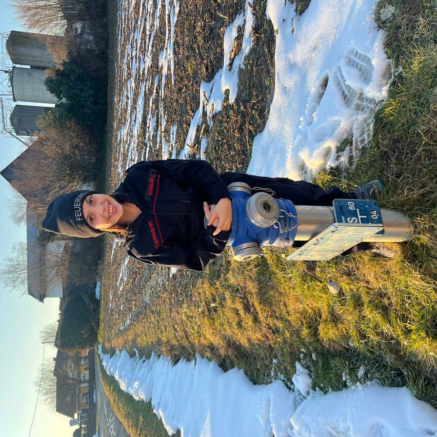 Eine Frau posiert mit einem blauen Wasserzähler auf einem Grashang mit Schnee. Ein Haus mit Schornstein und einem Speichertank ist im Hintergrund.