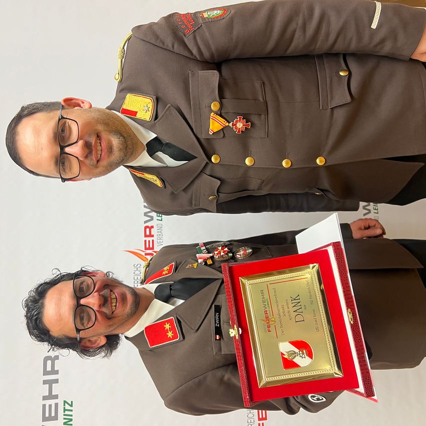 Zwei Männer in Militäruniformen lächeln und posieren für ein Foto. Der Mann vorne hält eine rote Plakette mit goldenen Akzenten. Hinter ihm steht ein anderer Mann mit Medaillen auf seiner Uniform.