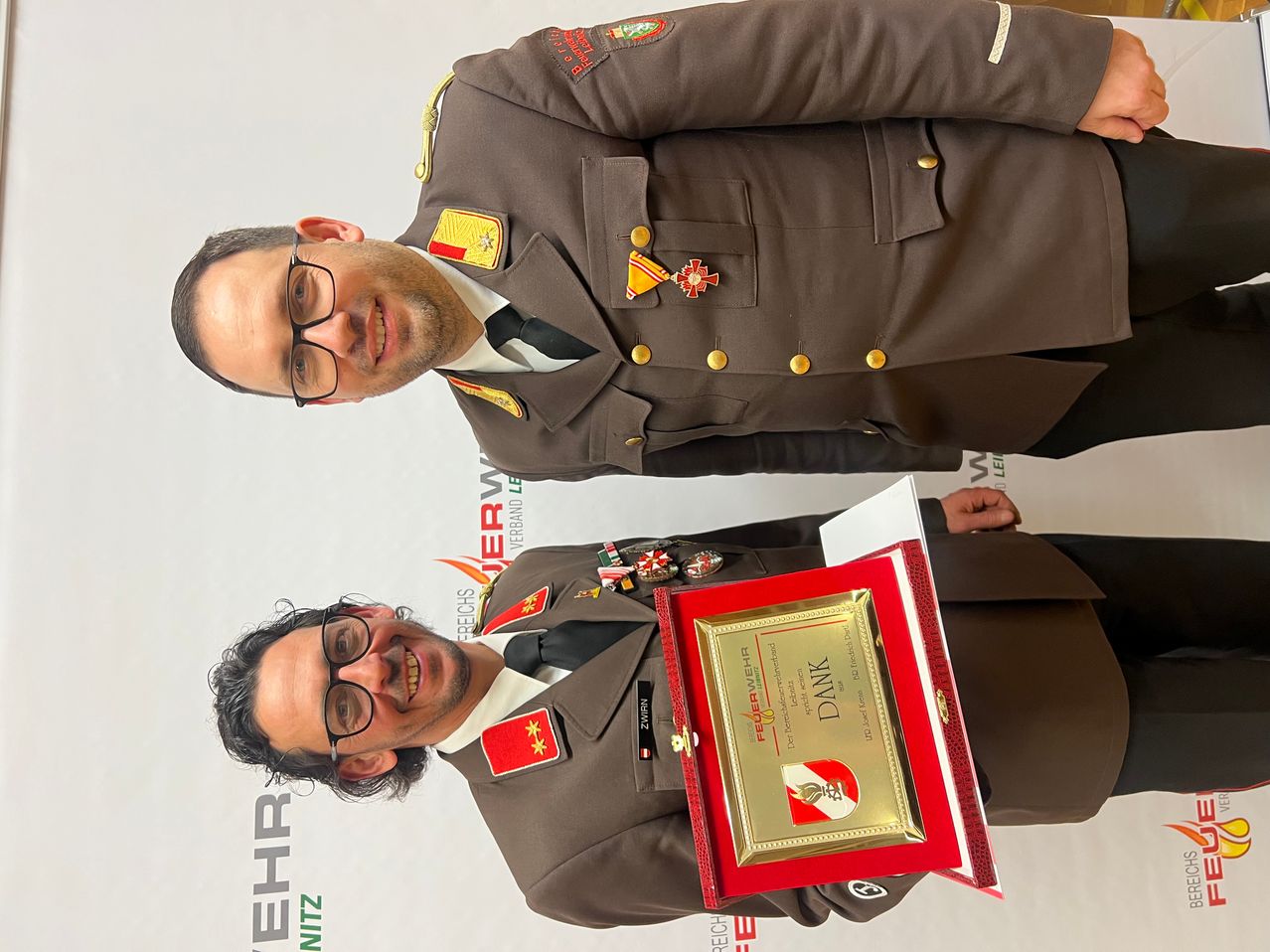 Zwei Männer in Militäruniformen lächeln und posieren für ein Foto. Der Mann vorne hält eine rote Plakette mit goldenen Akzenten. Hinter ihm steht ein anderer Mann mit Medaillen auf seiner Uniform.
