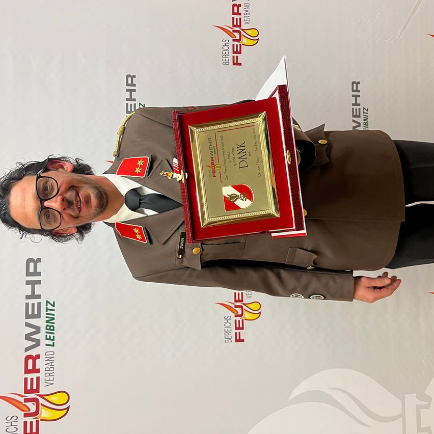 Ein Mann in Uniform hält eine rote Plakette mit goldenem Text und lächelt für die Kamera. Der Hintergrund zeigt Logos und Text, die mit einer Feuerwehr in Verbindung stehen.