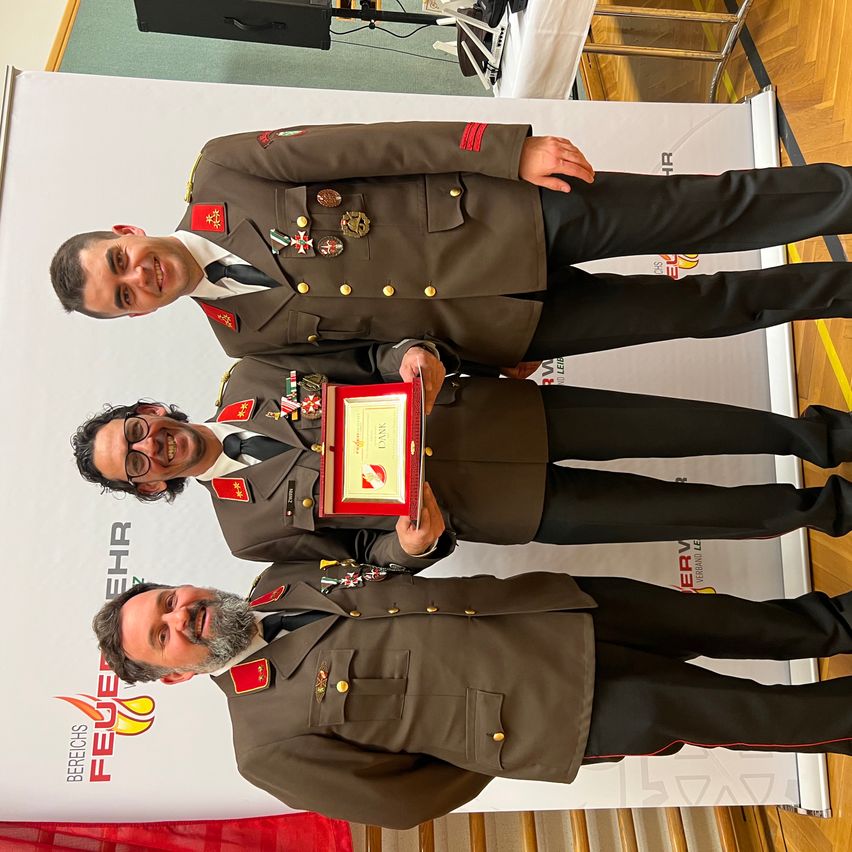 Drei Männer in Militäruniformen stehen zusammen und lächeln. Sie halten eine gerahmte Urkunde. Der Mann in der Mitte hält den Rahmen mit beiden Händen. Hinter ihnen ist ein Banner mit Text.