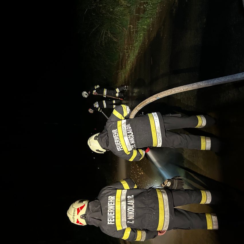Feuerwehrleute in Schutzkleidung löschen ein Feuer in einem dunklen Bereich. Ein Feuerwehrmann sprüht Wasser, während zwei andere mit einer Feuerwehrschläuche helfen.
