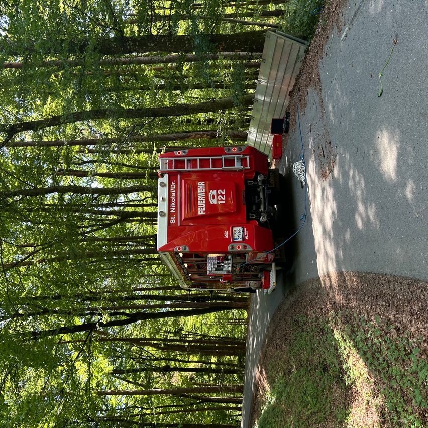 Ein roter Feuerwehrwagen steht auf der Seite einer Straße in einem Wald mit ausgefahrener Leiter und einer Schläuche auf dem Boden.