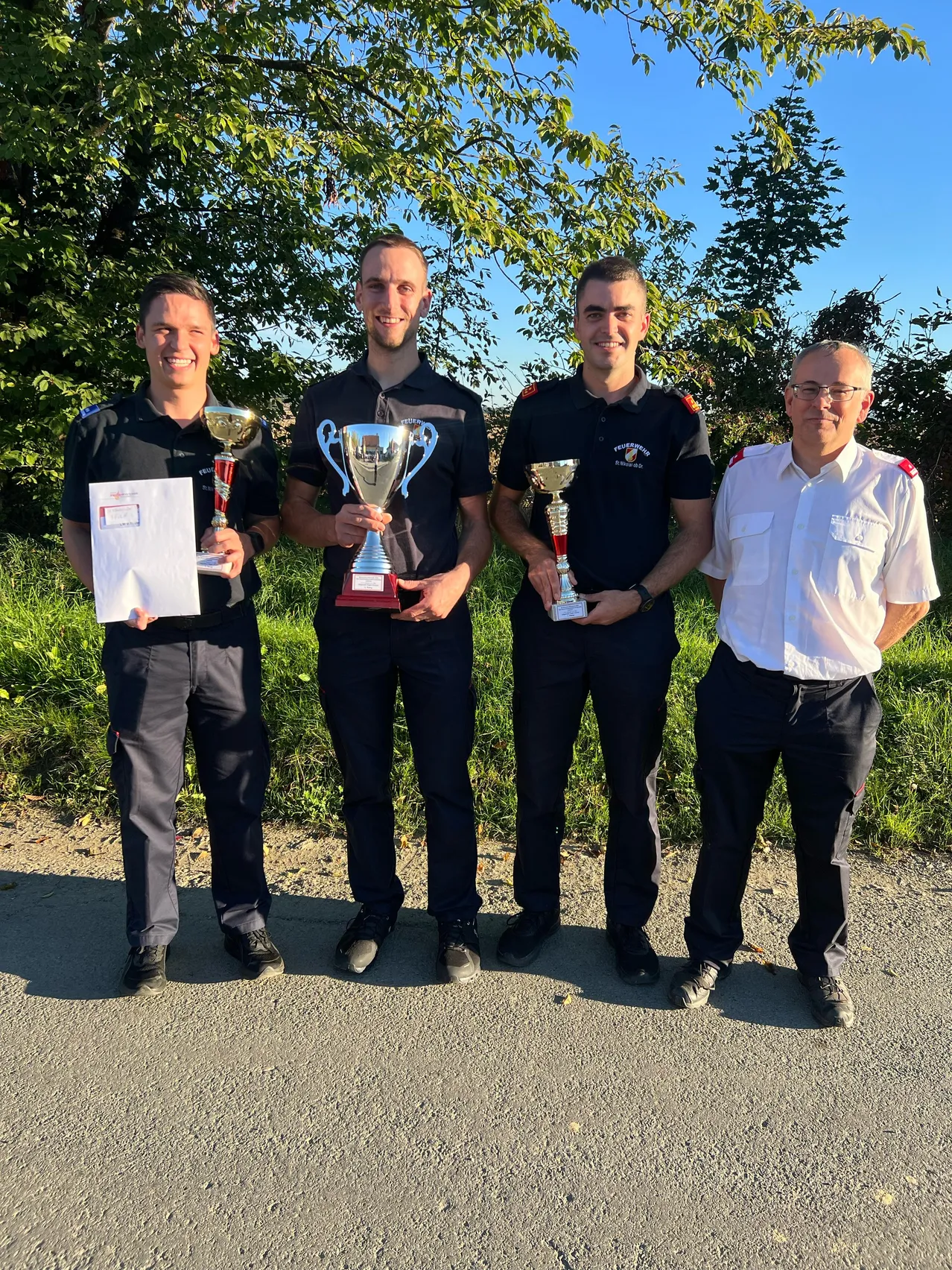 Vier Männer in Uniform, zwei mit Trophäen und einer mit einem Zertifikat, stehen auf einer Straße mit Bäumen im Hintergrund.