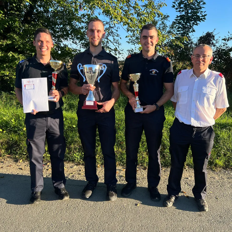 Vier Männer in Uniform, zwei mit Trophäen und einer mit einem Zertifikat, stehen auf einer Straße mit Bäumen im Hintergrund.