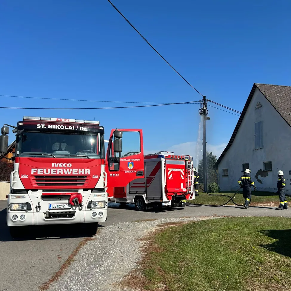 Ein Feuerwehrauto von St. Nikolai/DE ist am Straßenrand geparkt, während ein anderes Feuerwehrauto Wasser auf ein Gebäude sprüht.