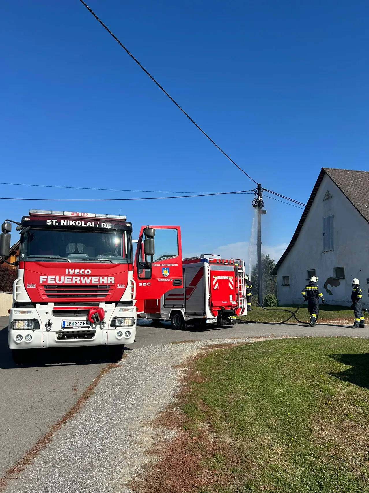 Ein Feuerwehrauto von St. Nikolai/DE ist am Straßenrand geparkt, während ein anderes Feuerwehrauto Wasser auf ein Gebäude sprüht.