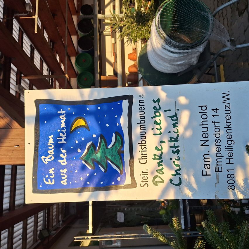 Ein Schild mit einer Weihnachtsbaumzeichnung und Text in einer Fremdsprache hängt im Freien. Das Schild hat einen blauen Hintergrund mit Schnee und einem Mond und ist an einer Holzstruktur montiert.