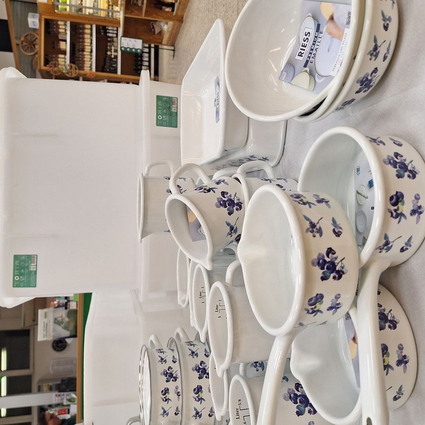 A collection of white ceramic cups and plates with blue floral patterns, displayed on a white surface. In the background, a wooden shelf with various items.