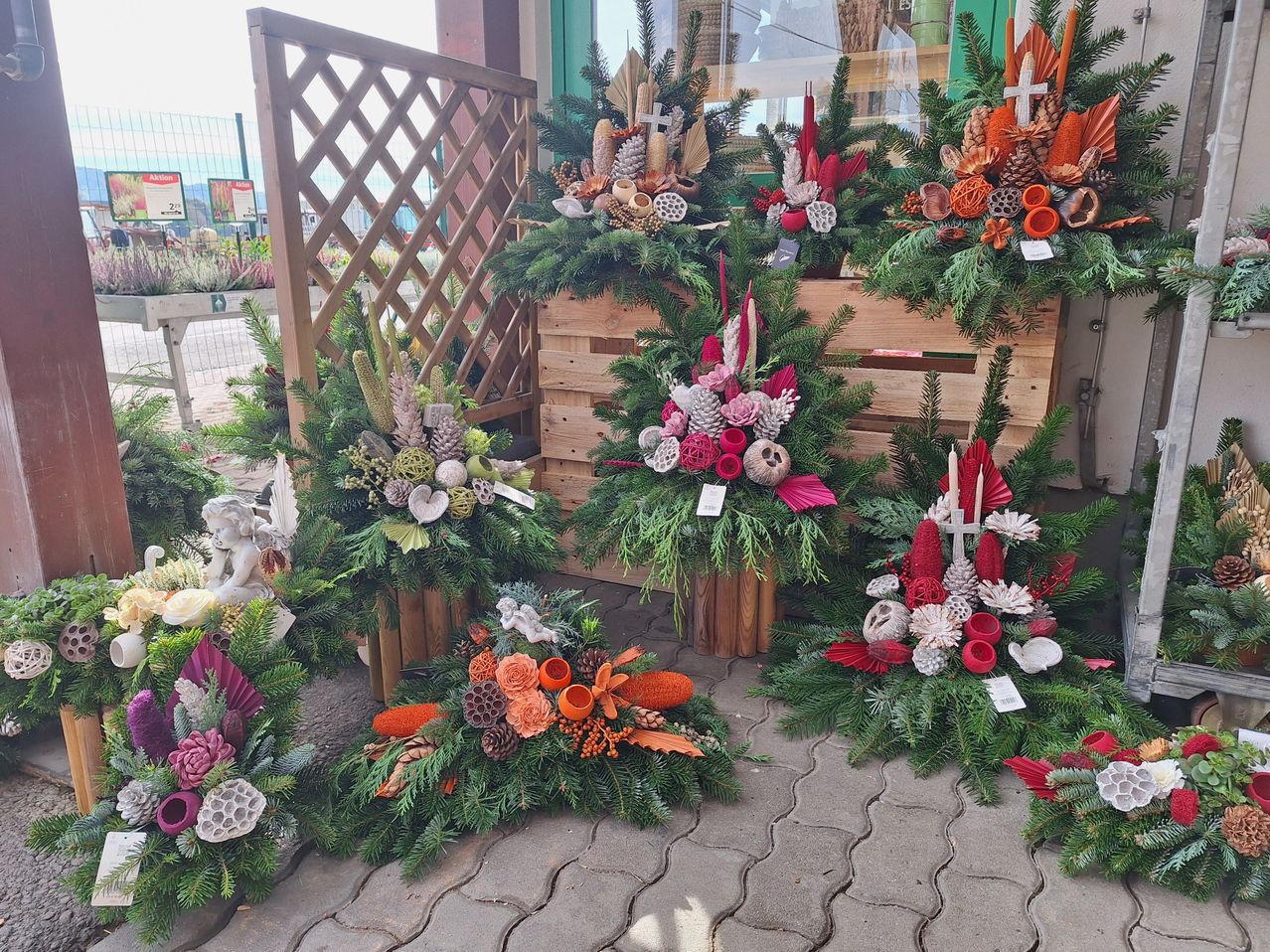Eine Außenausstellung von Kränzen aus Kiefern, Blumen und anderen Materialien, auf Holzständern und einer Holzkiste platziert, mit Kreuzen und Kerzen.