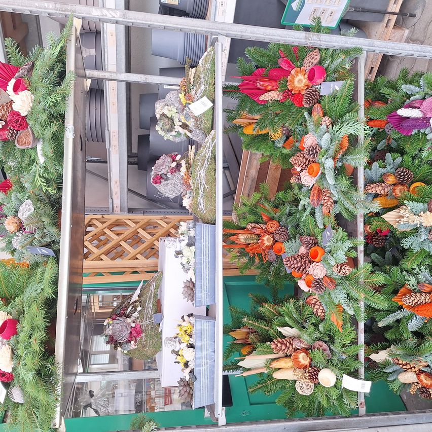 Eine Ausstellung verschiedener Kränze aus einer Kombination von frischen und künstlichen Blumen, Tannenzapfen und Grün. Die Kränze sind in einer Reihe auf einem Metallrahmen angeordnet.