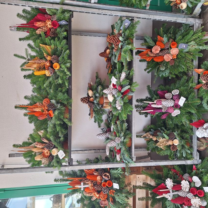 Ein vertikaler Blick auf mehrere Kränze, die an einem Metallrahmen hängen, geschmückt mit Tannenzapfen, Blättern und Ornamenten. Die Kränze variieren in Größe und Farbe, einige mit einem Kreuz.