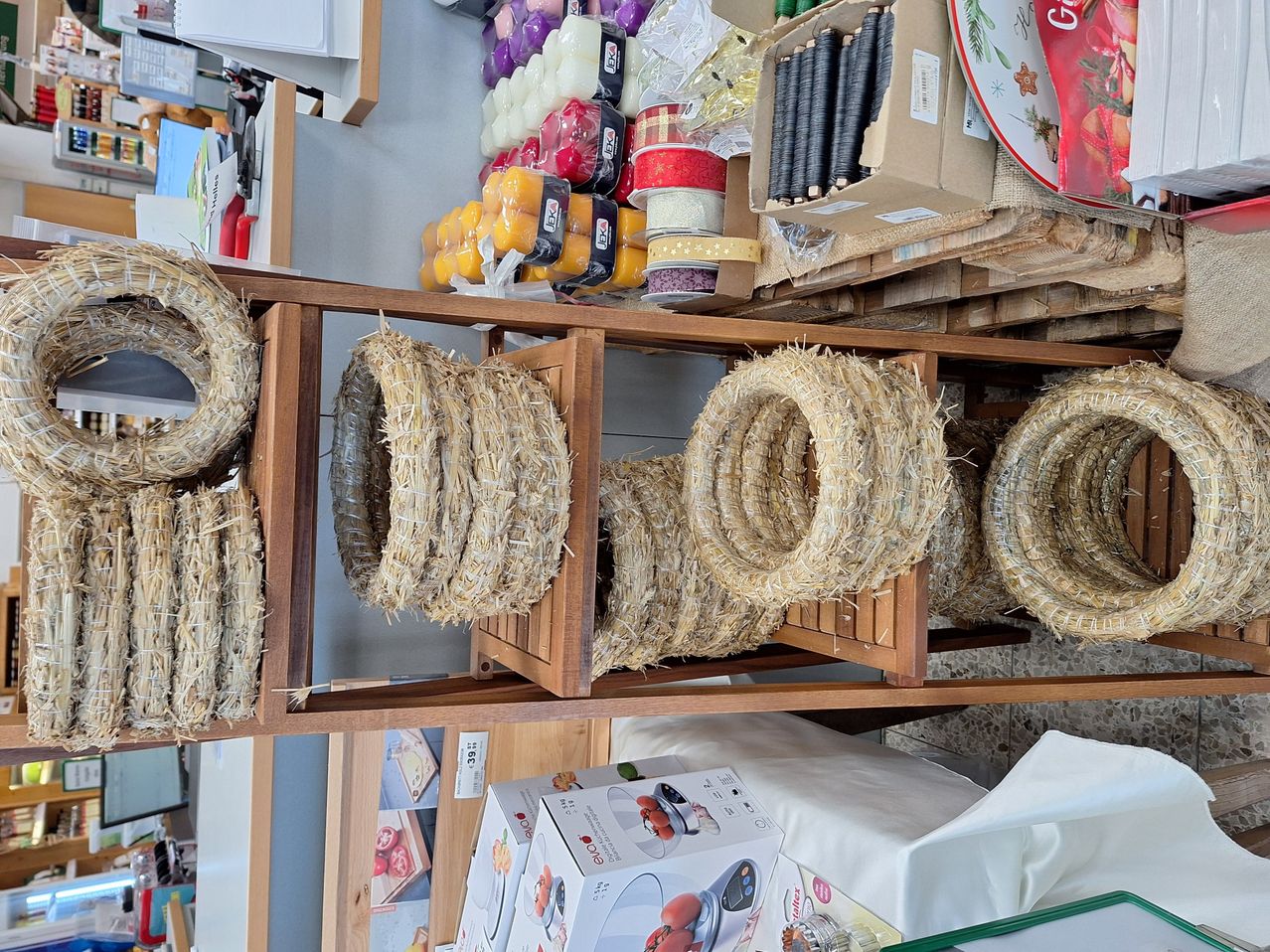 A wooden shelf displays several straw wreaths. The shelf also contains other items, including candles and ribbons. In the background, there are more boxes and items, suggesting a festive or decorative setting.